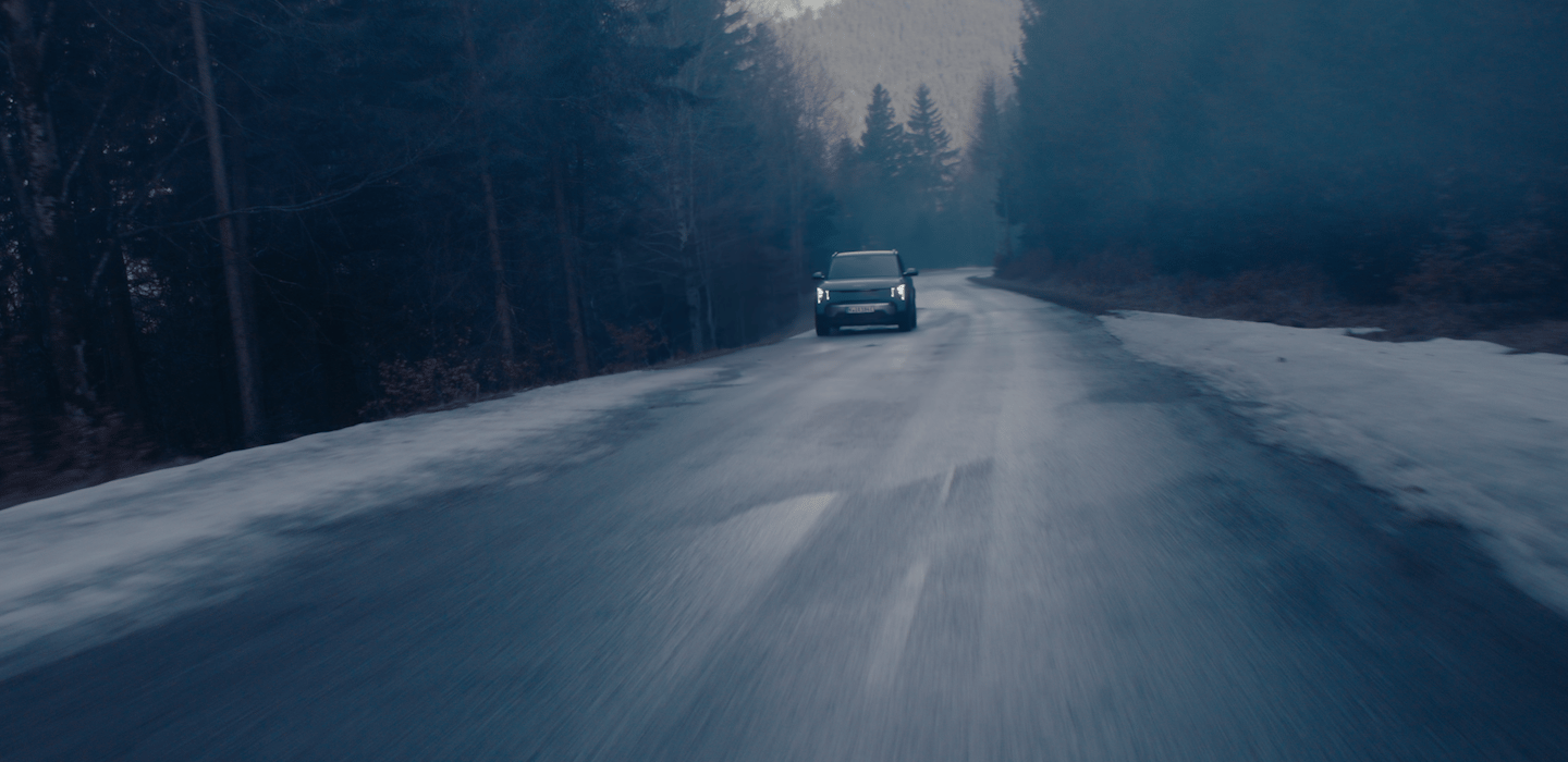 Dunkle, atmosphärische Aufnahme eines Kia Fahrzeugs, das bei Dämmerung mit eingeschalteten Scheinwerfern auf einer nassen Waldstraße auf die Kamera zufährt. Entlang der Straße liegen vereinzelt Schneereste.